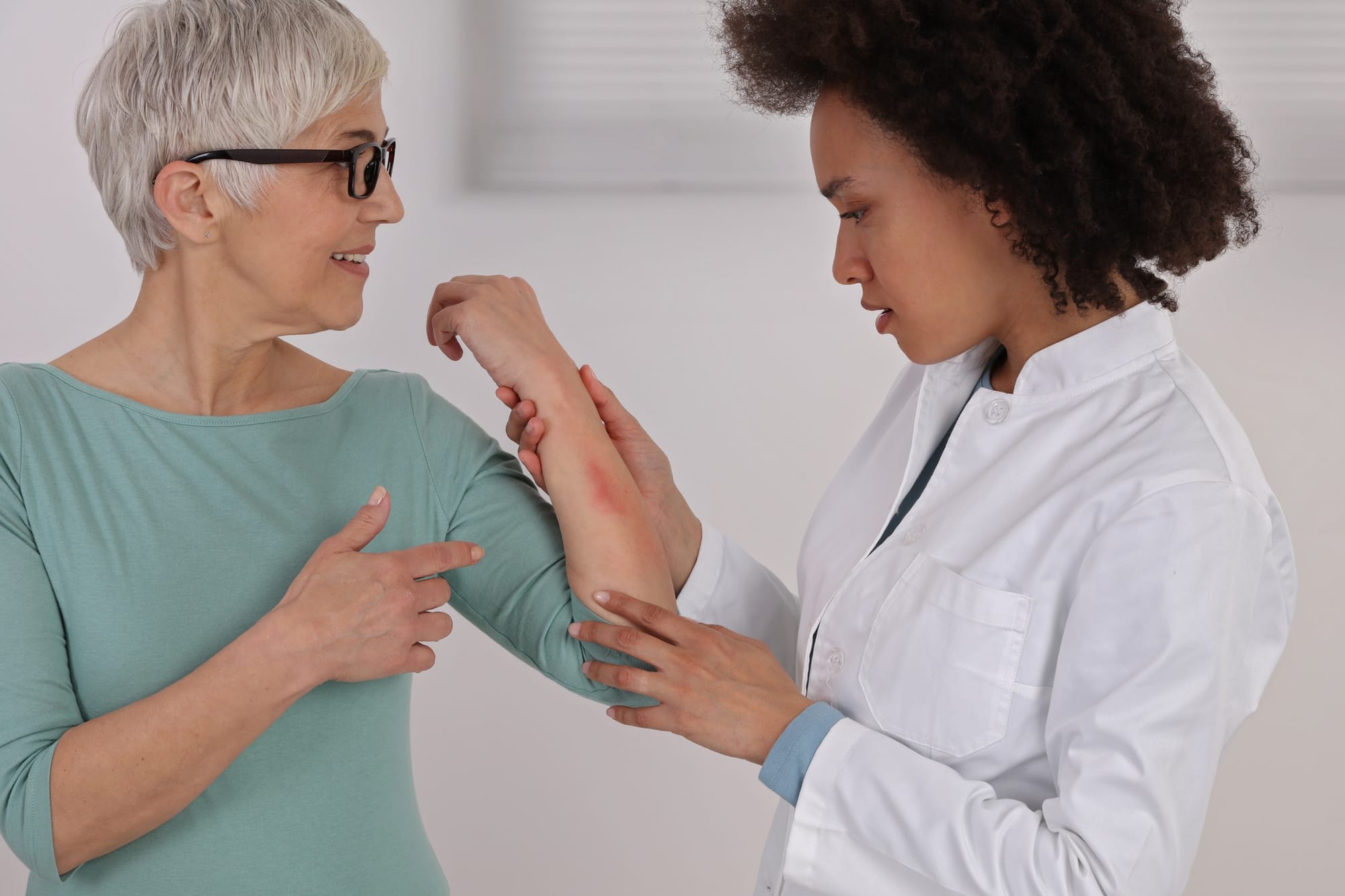 Stock photograph of models: a smiling grey-haired woman in a teal top shows a red patch on her forearm to a doctor in a white coat